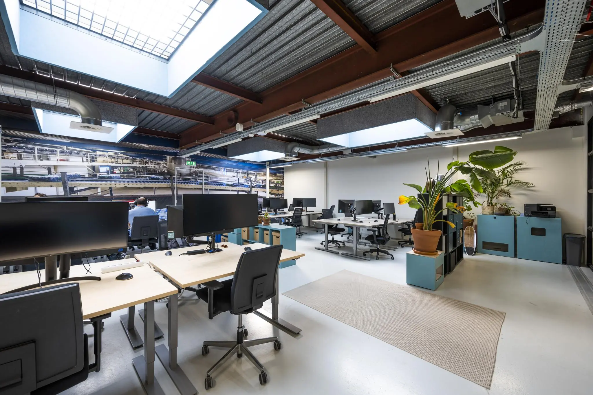 Modern, open kantoorinterieur aan de Joan Muyskenweg met bureaus, grote beeldschermen, planten en veel natuurlijk licht via dakramen.